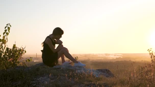 Ganska ledsen kvinna i svart kort sommar klänning sitter på en klippa tänkande utomhus vid solnedgången. Fashionabla kvinnliga överväger i varm kväll i naturen — Stockvideo
