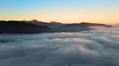 Ufuktaki dağ tepelerinin uzak karanlık siluetleri ile beyaz yoğun sis üzerinde renkli gün doğumunun havadan görüntüsü