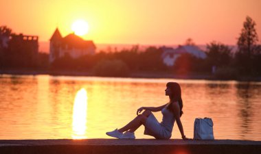 Sıcak bir akşamda göl kıyısında yalnız başına oturan yalnız bir kadın. Yalnızlık ve doğa konseptinde rahatlama.