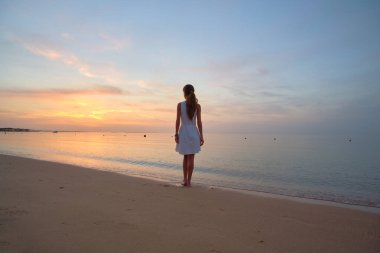Yalnız genç bir kadın kumsalda duruyor, deniz kenarında sıcak tropikal bir akşamın tadını çıkarıyor.