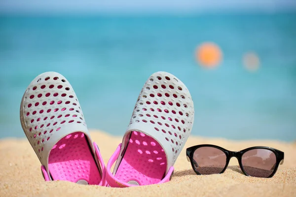 Sıcak, güneşli bir günde tropik deniz kıyısındaki kumlu kumsalda takunya ayakkabıları ve siyah koruyucu güneş gözlükleri. Yaz tatili kavramı