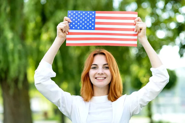 Szczęśliwa rudowłosa dziewczyna pozująca z flagą USA nad głową stojąca na świeżym powietrzu w letnim parku. Pozytywna młoda kobieta świętująca dzień niepodległości Stanów Zjednoczonych — Zdjęcie stockowe
