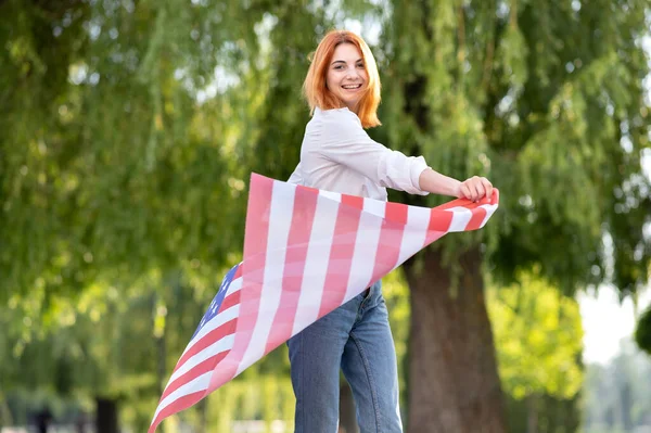 Jovem mulher ruiva feliz posando com bandeira nacional dos EUA de pé ao ar livre no parque de verão. Menina positiva celebrando o dia da independência dos Estados Unidos — Fotografia de Stock