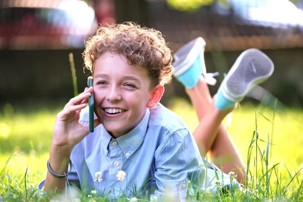 Šťastný dospívající chlapec mluví na mobilním telefonu venku v letním parku. Koncept přátelství online — Stock fotografie