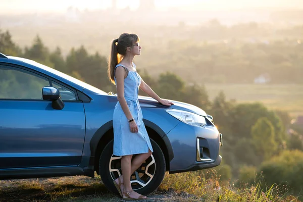 Mavi elbiseli mutlu genç kadın sürücü sıcak yaz akşamının tadını çıkarıyor arabasının yanında duruyor. Seyahat ve tatil kavramı