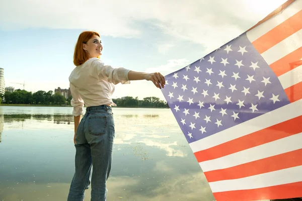 Joyeux jeune femme avec le drapeau national des États-Unis sur ses épaules avec des bâtiments de haute ville en arrière-plan célébrant la fête de l'indépendance des États-Unis — Photo