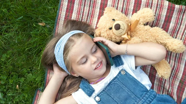 Lilla glada barn flicka som ligger på en filt på grön gräsmatta på sommaren med sin nalle talar på mobiltelefon — Stockfoto