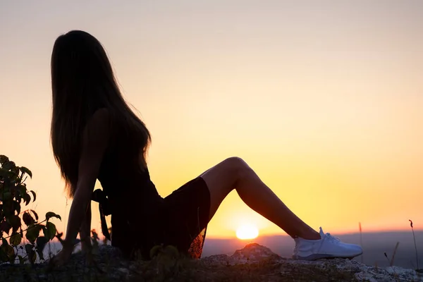 Young elegant woman in black short dress sitting on a rock relaxing outdoors at summer evening. Fashionable female enjoying warm sunset in nature — Stock Photo, Image