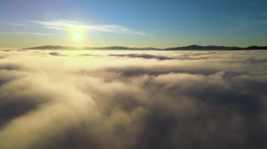 Beyaz yoğun bulutlar ve ufuktaki uzak dağların üzerinde canlı sarı gündoğumunun havadan görüntüsü.