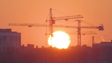 Gün batımında yüksek apartman binalarının inşaat sahasında büyük batan güneşli kule vinçlerinin koyu gölgesi. Gayrimenkul geliştirme