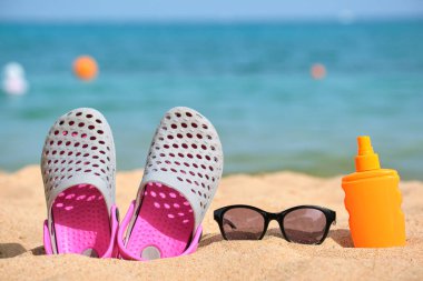 Sıcak güneşli bir günde, tropik deniz kıyısında takunya ayakkabıları, güneş kremi ve siyah koruyucu güneş gözlüklerine yakın çekim. Yaz tatili kavramı