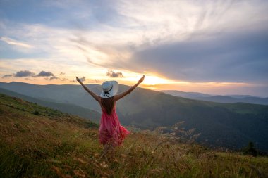 Kırmızı elbiseli mutlu bir kadın yürüyüşçünün arka görüntüsü çimenli bir tepede rüzgarlı bir sonbahar akşamında kollarını açıp güneşin batışının keyfini çıkarırken.