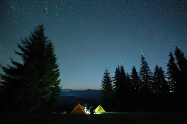 Yanlız yürüyüşçülerin silueti, karanlık dağlarda gece vakti gökyüzünün altında, aydınlık turist çadırlarının yanında şenlik ateşi yakmanın yanı sıra dinleniyor. Aktif yaşam tarzı ve açık hava yaşam konsepti