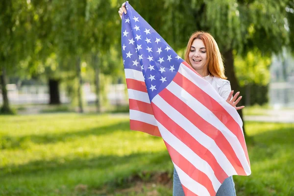 Jovem feliz posando com a bandeira nacional dos EUA de pé ao ar livre no parque de verão. Menina positiva com bandeira dos Estados Unidos ao ar livre — Fotografia de Stock