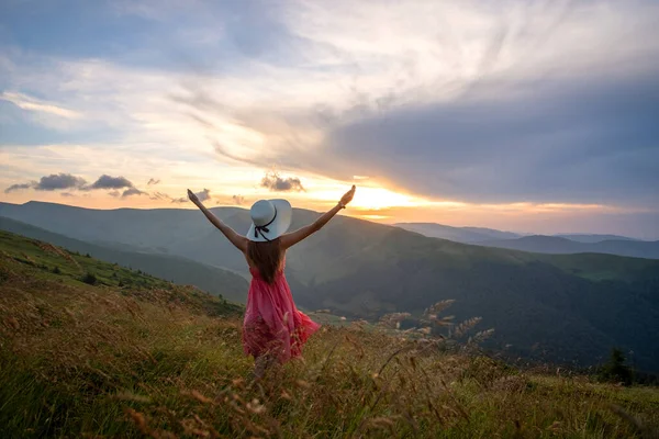 Visão traseira de uma mulher feliz caminhante em vestido vermelho em pé na colina gramada em uma noite ventosa em montanhas de outono com braços estendidos desfrutando de vista da natureza ao pôr do sol — Fotografia de Stock