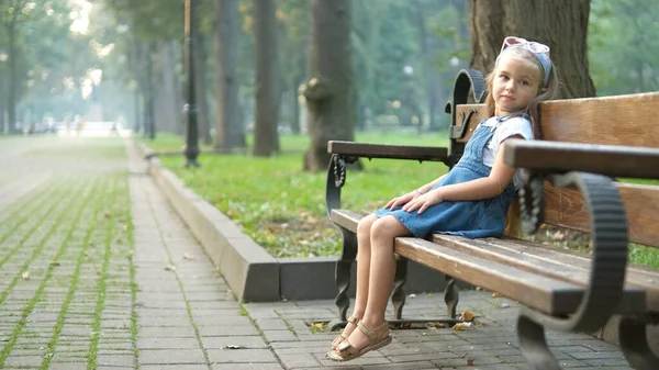 Pequeña niña feliz sentada en un banco descansando en el parque de verano —  Fotos de Stock