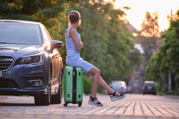 Joven mujer cansada con la maleta sentada al lado del coche esperando a alguien. Concepto de viajes y vacaciones — Foto de Stock