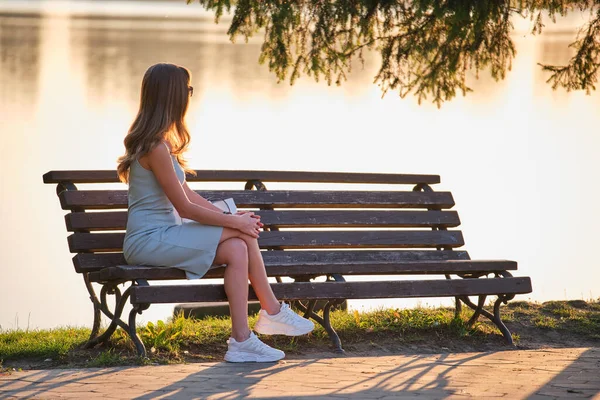 Eine junge Frau im Sommerkleid sitzt an einem warmen Abend entspannt auf einer Bank am See. Wellness abseits des alltäglichen Trubels — Stockfoto