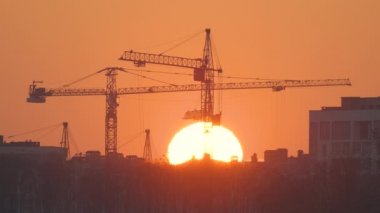 Gün batımında yüksek apartman binalarının inşaat sahasında büyük batan güneşli kule vinçlerinin koyu gölgesi. Gayrimenkul geliştirme