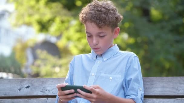 Jeune garçon jouant jeu sur son smartphone à l'extérieur dans le parc d'été. Addiction du concept de gadgets électroniques — Video