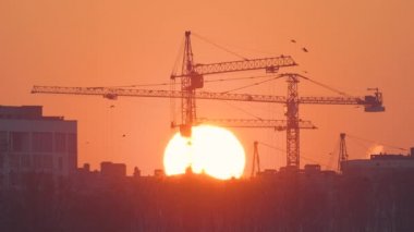 Gün batımında yüksek apartman binalarının inşaat sahasında büyük batan güneşli kule vinçlerinin koyu gölgesi. Gayrimenkul geliştirme