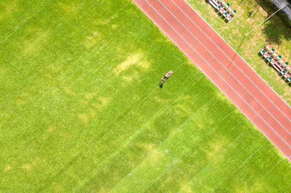 Vista aérea da pequena figura do trabalhador cortando grama verde com mashine de corte no campo de estádio de futebol com pistas de corrida vermelhas no verão — Fotografia de Stock