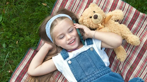 Lilla glada barn flicka som ligger på grön gräsmatta med sin nalle talar på mobiltelefon — Stockfoto