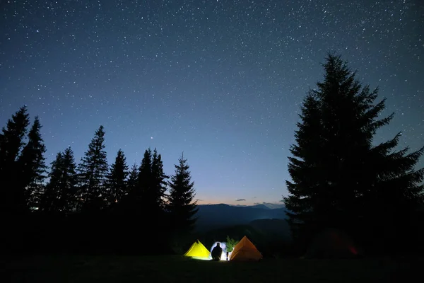 Silhouette de randonneur solitaire reposant à côté de feu de joie brûlant près des tentes touristiques éclairées sur le camping dans les montagnes sombres sous le ciel nocturne avec des étoiles. Style de vie actif et concept de vie en plein air — Photo