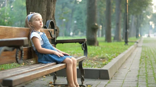 Malá unavená holčička sedící na lavičce se zavřenýma očima odpočívající v letním parku — Stock fotografie