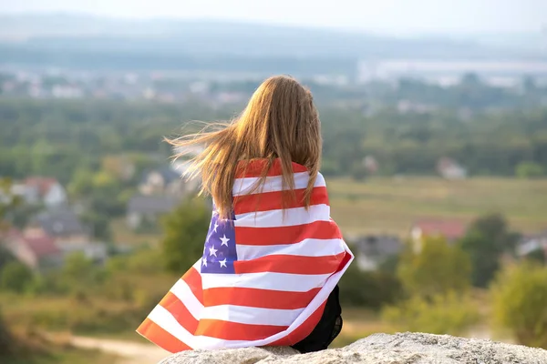 Young pretty american woman with long hair holding waving on wind USA flag on her sholders resting outdoors enjoying warm summer day — Stock Photo, Image