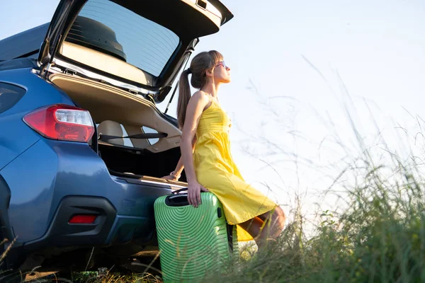 Mujer joven descansando en una maleta verde cerca de su coche en la naturaleza de verano. Concepto de viajes y vacaciones — Foto de Stock