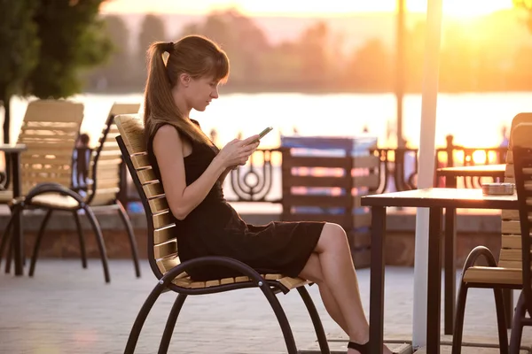 한 젊은 여성은 길거리 카페 테이블에 앉아 따뜻 한 여름날 저녁 야외에서 핸드폰을 검색 했습니다. 의사소통 과 이동 통신 개념. — 스톡 사진