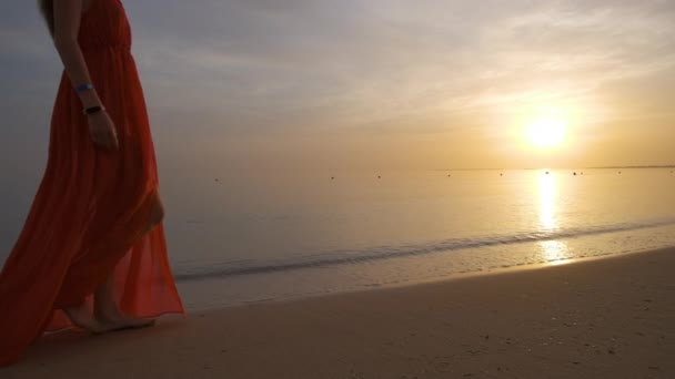 Jeune femme en robe rouge marchant seule sur une plage de sable au bord de la mer profitant d'une chaude soirée tropicale — Video