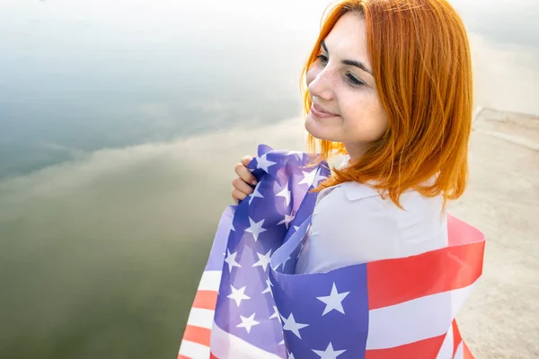 Szczęśliwa uśmiechnięta ruda dziewczyna z flagą USA na ramionach. Pozytywna młoda kobieta świętująca dzień niepodległości Stanów Zjednoczonych — Zdjęcie stockowe