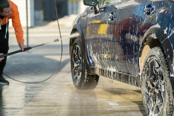 Młody kierowca myje samochód bezdotykowym strumieniem wody pod wysokim ciśnieniem w samoobsługowej myjni samochodowej — Zdjęcie stockowe