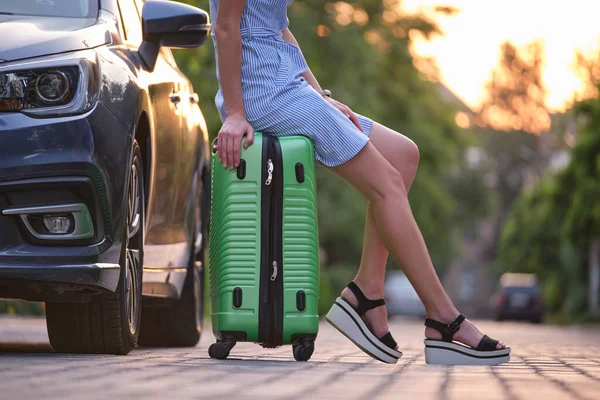 Großaufnahme junger weiblicher Beine, die auf einer Koffertasche neben dem Auto sitzen. Reise- und Urlaubskonzept — Stockfoto