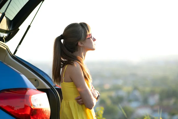 黄色のドレスを着た幸せな若い女性ドライバーは、彼女の車の横に立って暖かい夏の夜を楽しんでいます。旅行と休暇のコンセプト — ストック写真