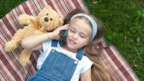 Lilla glada barn flicka som ligger på en filt på grön gräsmatta på sommaren med sin nalle talar på mobiltelefon — Stockfoto