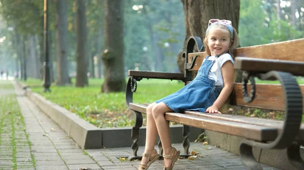 Mała szczęśliwa dziewczynka siedzi na ławce odpoczywając w letnim parku — Zdjęcie stockowe