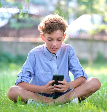 Yaz parkında akıllı telefonuyla oyun oynayan genç bir çocuk. Elektronik cihazlar konseptinden bağımlılık