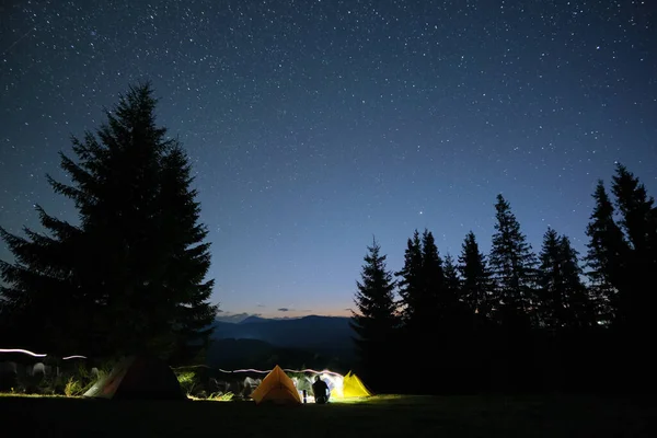 Silhouette de randonneur solitaire reposant à côté de feu de joie brûlant près des tentes touristiques éclairées sur le camping dans les montagnes sombres sous le ciel nocturne avec des étoiles. Style de vie actif et concept de vie en plein air — Photo