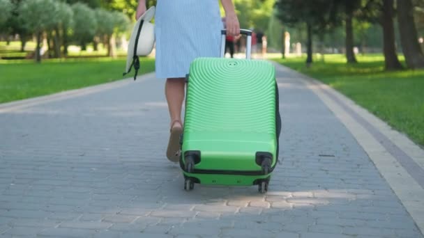 Jonge vrouw die op de stoep van het park loopt met groene koffer op zomerdag. Reizen en vakantie concept — Stockvideo