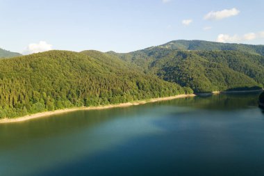 Yoğun yeşil ormanlarla kaplı yüksek dağ tepeleri arasında berrak mavi suyu olan büyük göl manzarası.