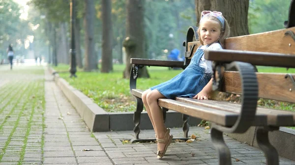 Malá šťastná holčička sedí na lavičce odpočívající v letním parku — Stock fotografie