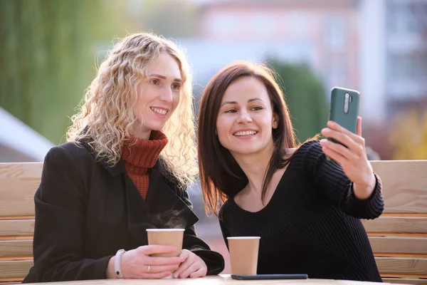 Duas mulheres felizes fazendo selfie com telefone celular sentado no café da rua da cidade. Amigos do sexo feminino curtindo tempo juntos ao ar livre — Fotografia de Stock