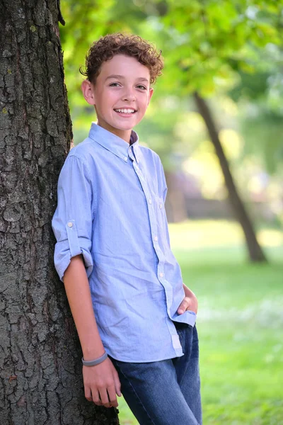 Молодий щасливий хлопчик розслабляється в літньому парку, спираючись на стовбур дерева. Концепція добробуту дітей — стокове фото