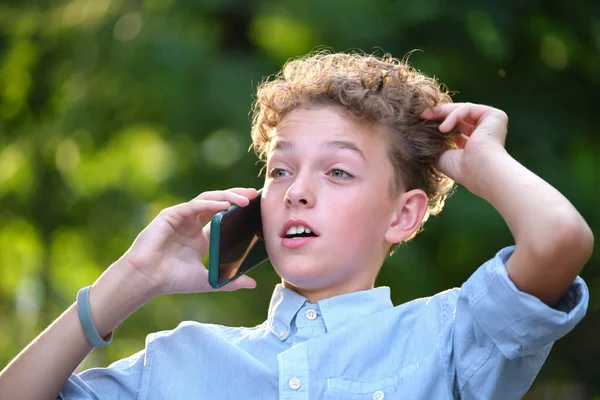 Mladý zmatený chlapec mluví na mobilní telefon venku v letním parku — Stock fotografie