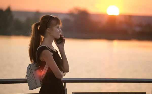 Молодая женщина, стоящая на берегу озера в летнем парке и разговаривающая по мобильному телефону на улице теплым вечером. Концепция связи и мобильной связи — стоковое фото