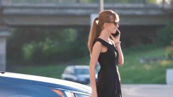 Νεαρή γυναίκα οδηγός στέκεται κοντά στο αυτοκίνητό της μιλώντας στο κινητό τηλέφωνο σε ένα δρόμο της πόλης το καλοκαίρι — Αρχείο Βίντεο