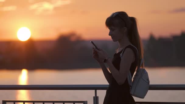Šťastná mladá žena relaxuje na břehu jezera v letním parku a mluví na svém mobilním telefonu venku za teplého večera. Koncept komunikace a mobilního připojení — Stock video